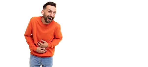 Ung man i orange tröja skrattar håller magen med händerna på en vit bakgrund — Stockfoto