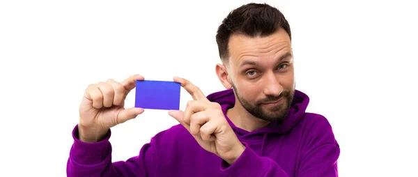 Un giovane con la barba con una felpa viola dimostra un biglietto da visita tra le mani su sfondo bianco — Foto Stock