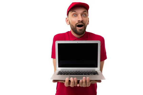 Mensajero de entrega sorprendido en uniforme rojo sosteniendo un ordenador portátil con la pantalla hacia adelante en su cara sorpresa y deleite —  Fotos de Stock