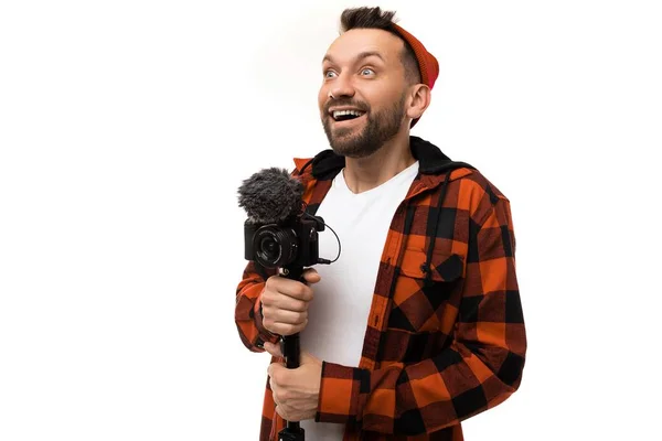 Inspirerad man hipster bloggare I en rutig röd skjorta och röd mössa med en kamera och en mikrofon i händerna ser upp till vänster med glada känslor i ansiktet — Stockfoto