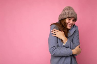 Güzel, pozitif, neşeli, neşeli, esmer kadının fotoğrafı. Pembe arka plan duvarında izole edilmiş, gri sonbahar ceketi ve gri, sıcak şapkalı, kameraya bakan ve soğuk hisseden bir kadın.
