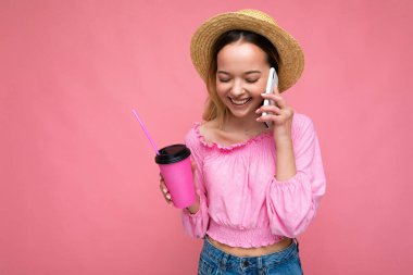 Pembe arka planda kahve içerken cep telefonuyla konuşan mutlu genç bir kadın.