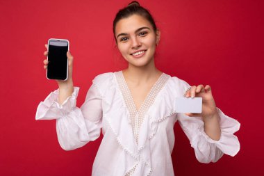 Resmî giysiler içinde pozitif neşeli bir kadının portresi ve cep telefonunu boş ekranla gösterme ve kırmızı arka planda izole edilmiş kameraya bakarak kesilen kredi kartı.