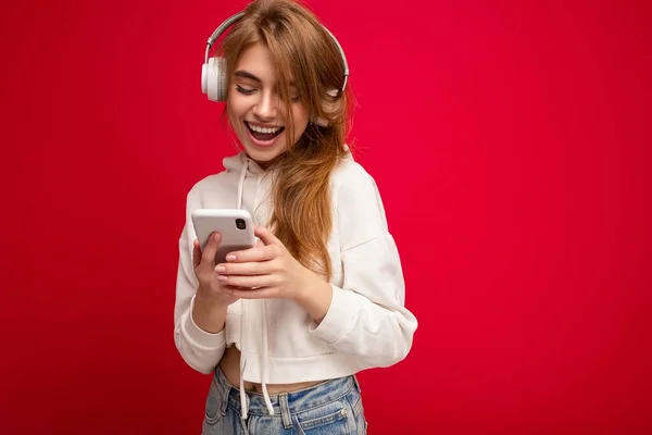 Photo of beautiful happy smiling young blonde woman wearing white hoodie isolated over the colourful background wall with copy space for text wearing white wireless bluetooth earphones listening to cool — Zdjęcie stockowe