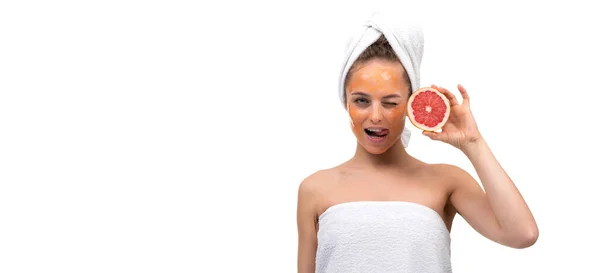Duştan sonra beyaz arka plandaki bir kadın bir gözünü kırpıyor ve yüzünde kozmetik bir maskeyle elinde kırmızı bir greyfurt tutuyor. — Stok fotoğraf