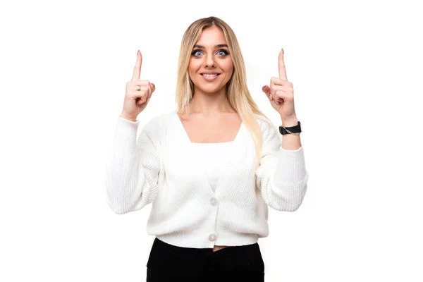 Φωτογραφία από μια όμορφη θετική ευτυχισμένη χαμογελαστή νεαρή ξανθιά γυναίκα με μακιγιάζ σε ένα κομψό λευκό πλεκτό πουλόβερ και μαύρο παντελόνι δείχνει τα δάχτυλα επάνω απομονώνονται σε ένα λευκό φόντο με κενό χώρο για κείμενο — Φωτογραφία Αρχείου