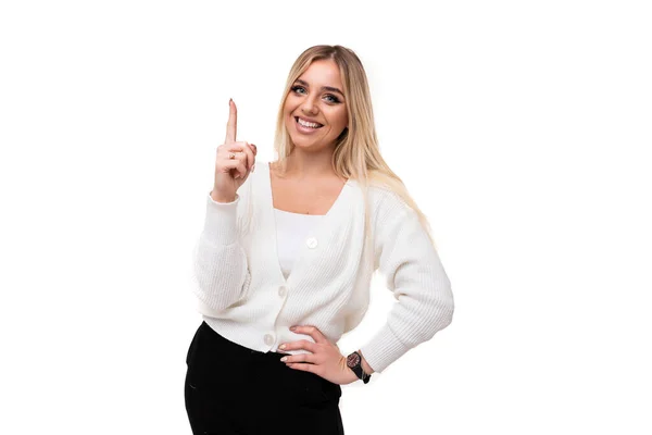 Foto van een mooie vrij positieve glimlach vrolijke jonge blonde vrouw met make-up in een stijlvolle witte gebreide trui en zwarte broek toont vinger omhoog geïsoleerd op een witte achtergrond met lege ruimte voor — Stockfoto