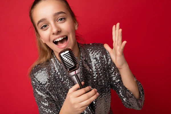 Mooi emotioneel verbaasd positief brunette klein meisje dragen stijlvolle glans jurk geïsoleerd over rode achtergrond muur zingen van het lied in de zilveren microfoon op zoek naar camera — Stockfoto