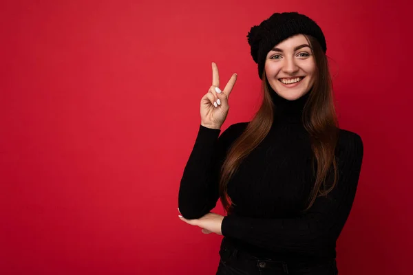 Фото молодой позитивной привлекательной брюнетки с искренними эмоциями в черном свитере и черной шляпе на красном фоне с копировальным пространством и жестом мира — стоковое фото
