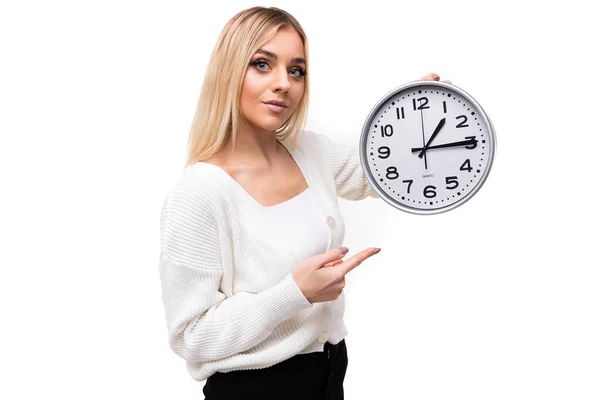 Foto seorang wanita cantik berambut pirang muda yang positif dengan sweater rajutan putih dan celana hitam menunjukkan jarinya pada jam besar yang terisolasi pada latar belakang putih dengan ruang fotokopi untuk teks — Stok Foto