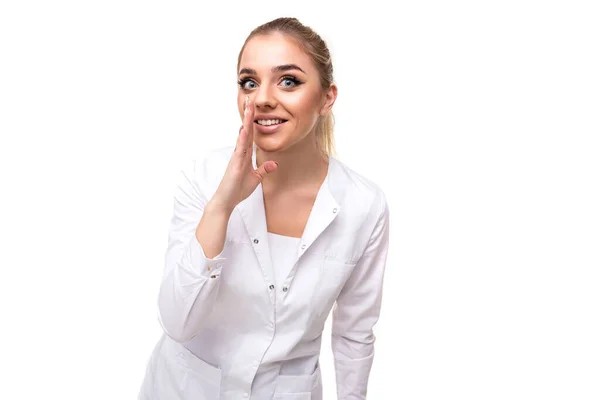 Photo of a beautiful pretty cute attractive positive young blonde woman with a ponytail and makeup in a white medical coat of a nurse tells a secret isolated on a white background with empty space for — Stock Photo, Image