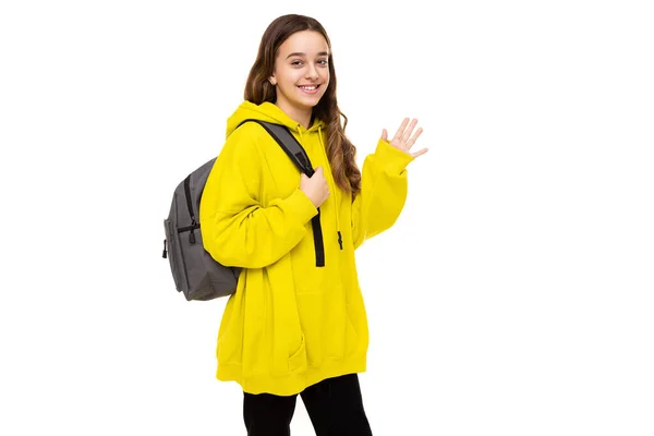 Photo of beautiful adorable cute smiling happy brunette teenage girl in stylish yellow sports hoodie with gray backpack waving hand isolated on white background with free space for text — 图库照片