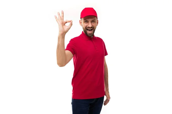 Kurýr v červených šatech na bílém pozadí ukazuje gesto symbolu Dobře, dívá se na kameru s úsměvem — Stock fotografie