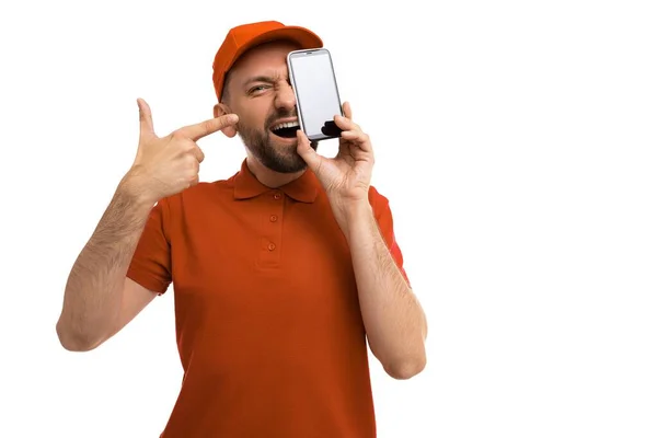 The courier demonstrates the application for ordering delivery on the smartphone screen, pointing at it with his finger, holds the phone at head level on a white background in a red uniform — 图库照片