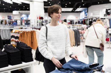 Gayri resmi ve şık beyaz kazaklı kısa saçlı güzel ve çekici bir esmer kadın alışveriş merkezindeki bir mağazadan şık kot pantolon ve kot elbise seçiyor. Dükkan konsepti