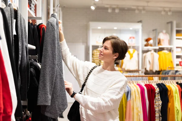 Photo of young attractive brunette woman with a short haircut in a white sweater chooses stylish and casual clothes in a store in a shopping mall. Shop concept — Stock Photo, Image