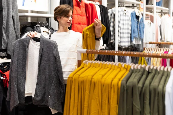 Photo of young attractive brunette woman with a short haircut in a white sweater chooses stylish and casual clothes in a store in a shopping mall. Shop concept — Stock Fotó