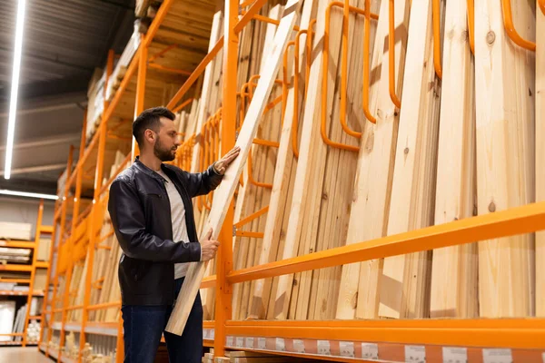 A man in a construction hypermarket in the lumber department holds a board in his hands next to a rack with wooden bars and clapboard for wall and facade decoration — Stock fotografie