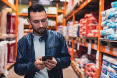 Hırdavatçıdaki inşaat malzemeleri arasında akıllı telefondan yazı yazan bir adam.