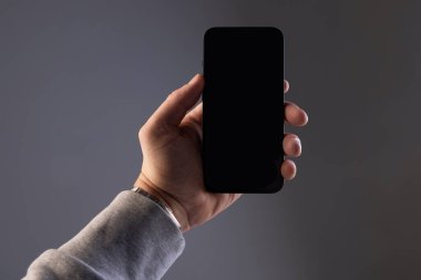 Gri eşofmanlı bir erkek eli, gri izole bir arkaplanda siyah dikdörtgen modern bir akıllı telefon tutar. Kopya-uzay için alanı olan bir ekran.