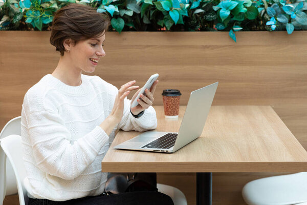 Молодая красивая женщина сидит в торговом центре за столом и работает за компьютером ноутбуком, используя мобильный телефон. Концепция фриланса и бизнеса