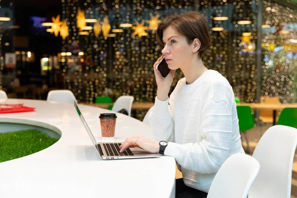 Joven mujer atractiva sentada en el patio de comidas de un centro comercial en una mesa y trabajando en un ordenador portátil. Café interior. Concepto independiente — Foto de Stock