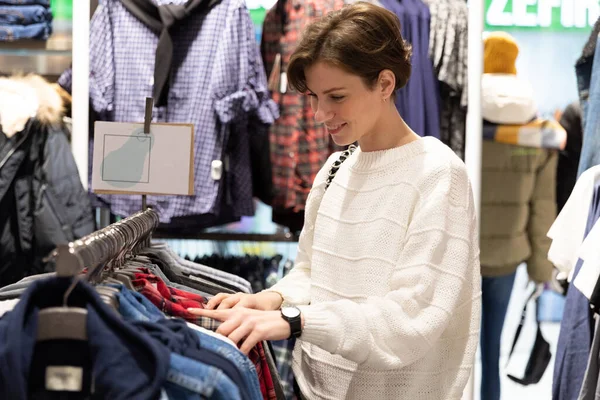 Young beautiful brunette woman with a short haircut in a white sweater chooses stylish clothes in a store in a shopping mall — Stock Photo, Image