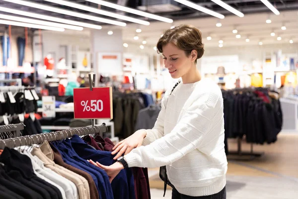 Young beautiful brunette woman with a short haircut in a white sweater chooses stylish clothes in a store in a shopping mall — Stock Photo, Image