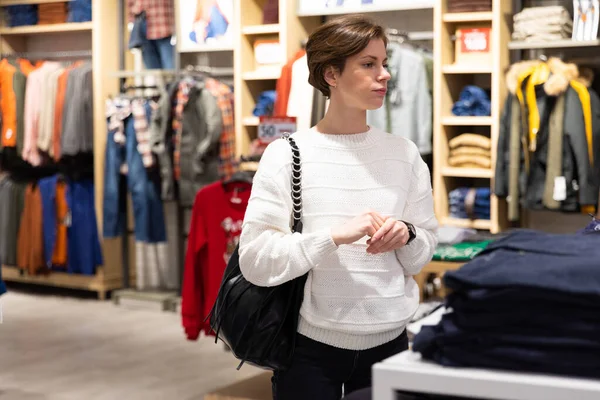 Young beautiful brunette woman with a short haircut in a white sweater chooses stylish clothes in a store in a shopping mall — Stock Fotó