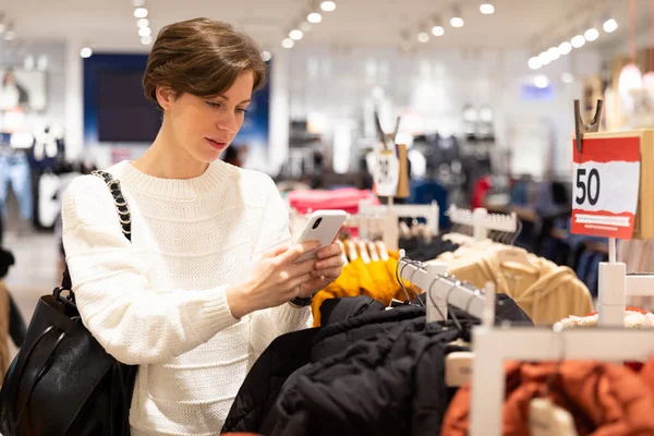 Fénykép a fiatal vonzó barna nő egy rövid frizura egy fehér pulóver választja elegáns és alkalmi ruhát egy boltban egy bevásárló központban. Üzletkoncepció — Stock Fotó