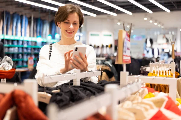 Adulto atraente mulher morena com um corte de cabelo curto em uma camisola branca escolhe roupas elegantes e casuais em uma loja em um centro comercial usando telefone celular — Fotografia de Stock