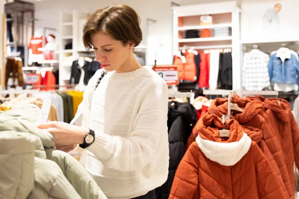 Photo of young attractive brunette woman with a short haircut in a white sweater chooses stylish and casual clothes in a store in a shopping centre — Stock Photo, Image