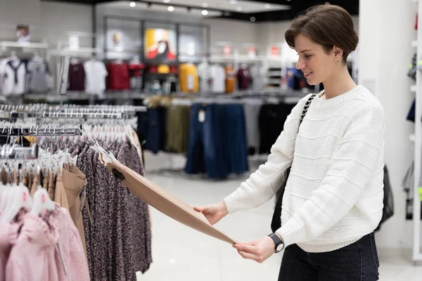 Egy fiatal, gyönyörű és vonzó barna nő, rövid hajjal, alkalmi és elegáns fehér pulóverben divatos ruhákat választ egy boltban egy bevásárlóközpontban. Üzletkoncepció — Stock Fotó