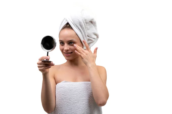 Belle femme d'âge moyen avec une peau bien soignée applique de la crème sur son visage et regarde dans un petit miroir sur un fond blanc — Photo