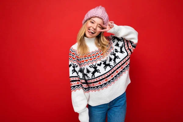Foto de joven positiva feliz hermosa rubia con emociones sinceras usando sombrero de punto rosa y jersey de invierno aislado sobre fondo rojo con espacio vacío — Foto de Stock