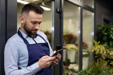 Çiçekçi dükkanında müdürün elinde bir telefonla buzdolabının yanında buketler var.