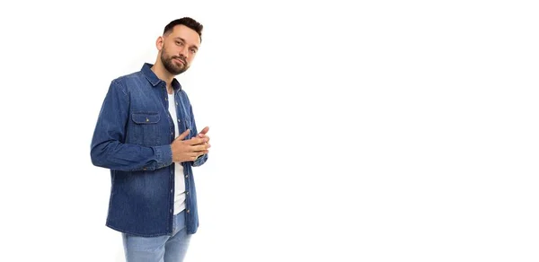 Мужчина средних лет с бородой в синей джинсовой рубашке и белыми футболками на белом фоне задумчиво смотрит в камеру — стоковое фото