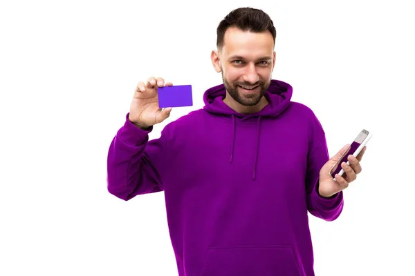 Moda elegantemente vestido homem de meia-idade com uma barba em uma camisola roxa demonstra um cartão bancário e uma aplicação em um smartphone — Fotografia de Stock