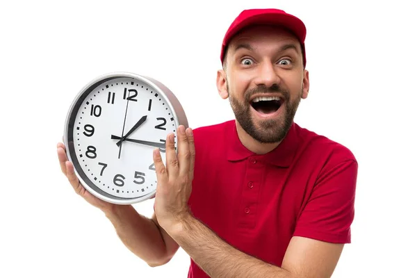 Correio carismático segurando um relógio com um sorriso no rosto em uma camiseta vermelha e um boné em um fundo branco, o conceito de entrega rápida e gerenciamento de tempo — Fotografia de Stock