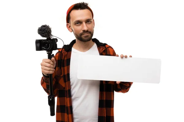 Мужчина-блогер в белой футболке и рубашке в красной и черной клетке с видеокамерой и микрофоном смотрит в объектив и держит табличку с пространством для копирования — стоковое фото