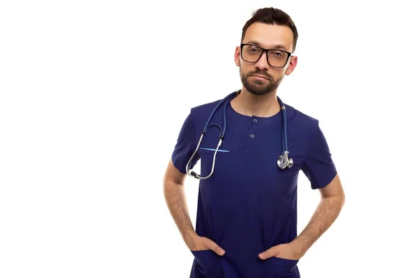 Een mannelijke arts met een bril op een witte achtergrond in blauwe kleren met een stethoscoop om zijn nek houdt zijn handen in zijn zakken en kijkt serieus in de camera — Stockfoto