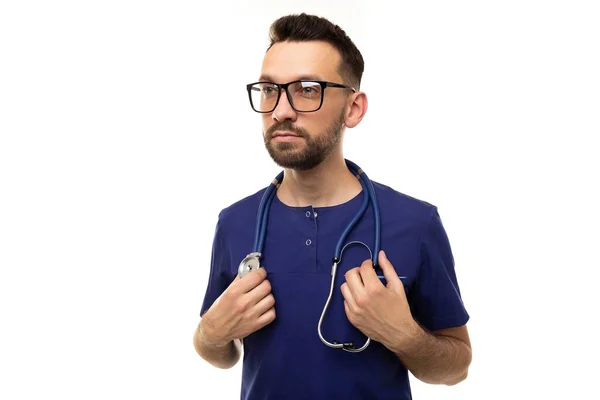 Een serieuze arts in bril en blauw uniform houdt een stethoscoop, kijkt naar links — Stockfoto