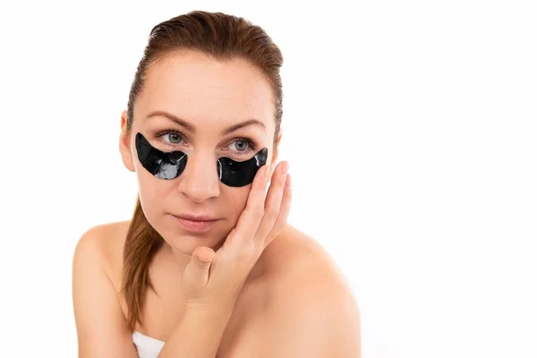 A woman applies cosmetic black patches under her eyes against a white wall — Stock Photo, Image