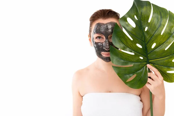 A young woman with a black cosmetic mask on her face made of activated charcoal in a towel after a shower on a white background peeps out from behind a leaf of this palm tree in her hands — Stock Photo, Image