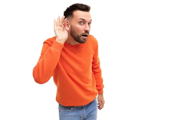 Un hombre en un suéter naranja escucha con la mano a la oreja sobre un fondo blanco —  Fotos de Stock
