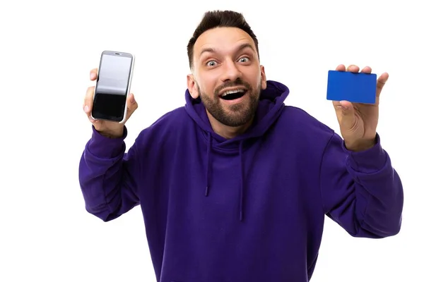 Homem carismático estranho com um sorriso e uma barba em uma camisola azul segurando um cartão de desconto e uma tela de telefone — Fotografia de Stock