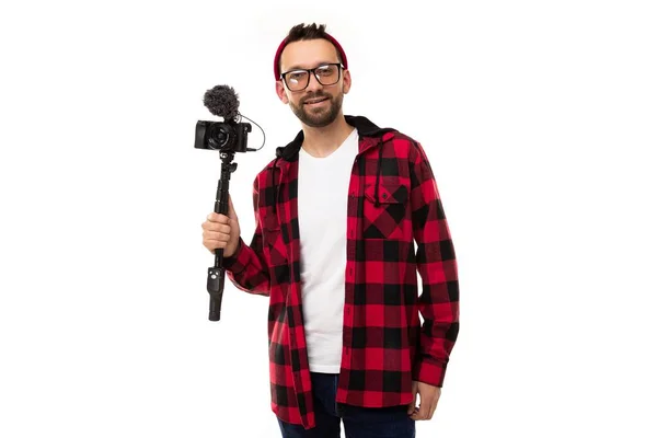 Ένας νεαρός blogger σε λευκό φόντο με καρό πουκάμισο και γυαλιά για την όραση κρατά μια κάμερα με ένα μικρόφωνο στα χέρια του — Φωτογραφία Αρχείου