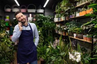 Bahçe merkezi müdürü, saksı bitkileriyle rafların önünde telefonla konuşuyor.