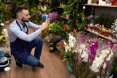 Adamın biri çiçekçide bir saksı bitkisinin akıllı telefonuyla fotoğraf çekiyor.
