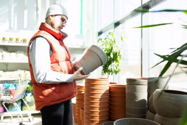 Elinde seramik bahçe tenceresi olan bir müşteri, saksılarla dolu bir rafın yanında.